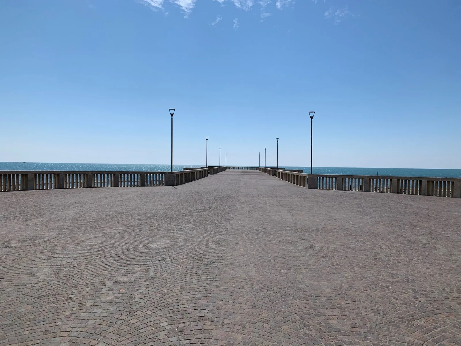 Il Pontile di Ostia in una giornata limpida: la piazza lastricata a sampietrini, le balaustre chiare e i lampioni che accompagnano la passerella verso il mare blu