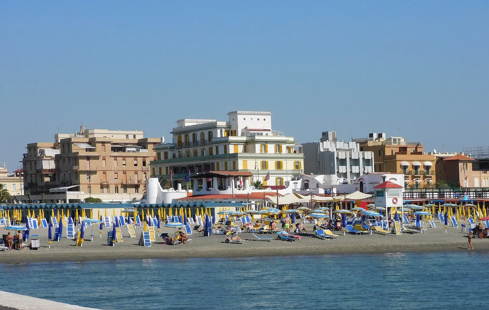 La spiaggia di Lido di Ostia d'estate vista dal mare, con file di ombrelloni gialli e blu, lettini, una torretta di salvataggio e i palazzi del lungomare sullo sfondo