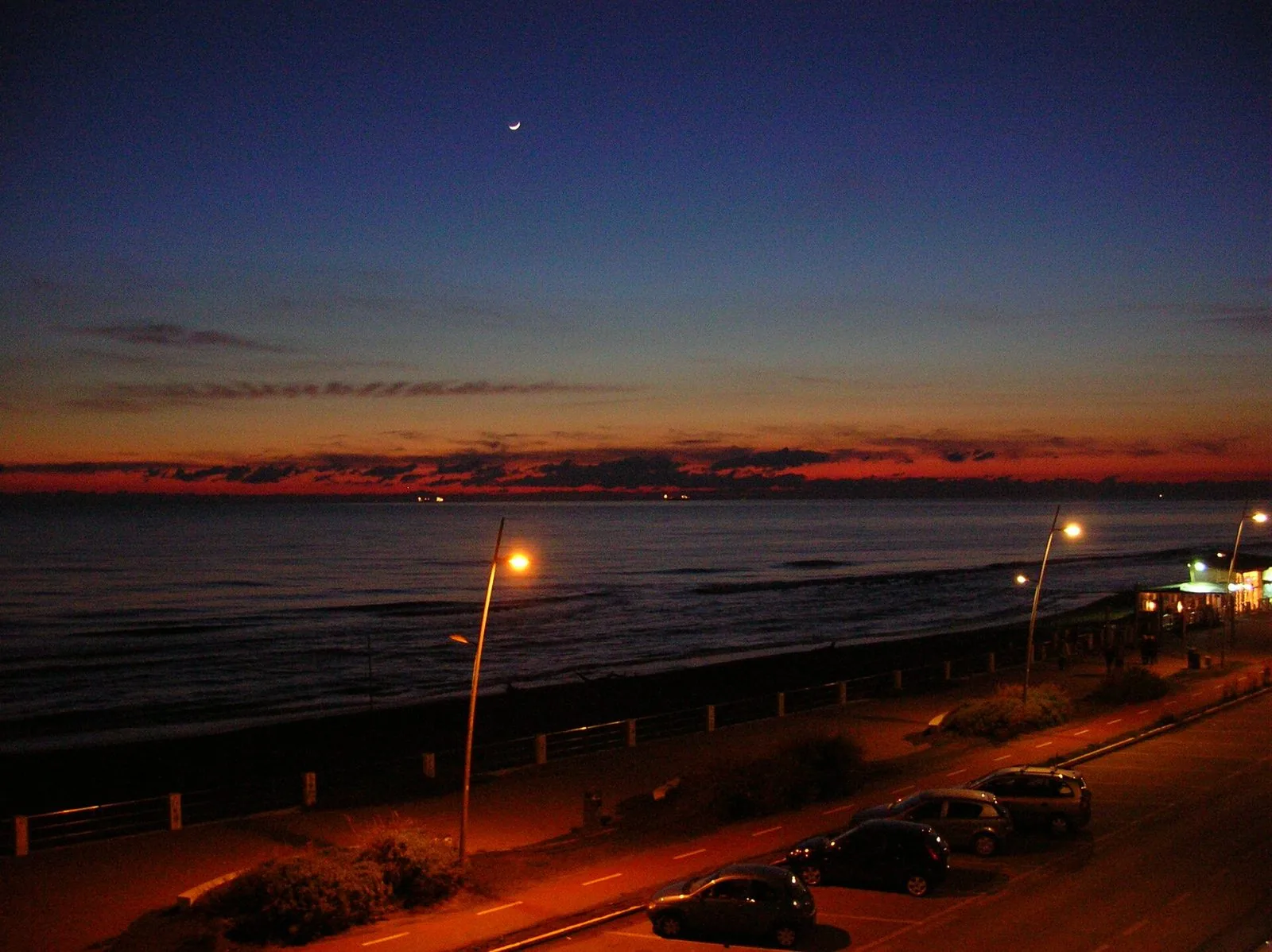 Il lungomare di Ostia al crepuscolo d'inverno, con il mare scuro, i lampioni accesi lungo la passeggiata e la pista ciclabile, il cielo blu con una sottile falce di luna e una striscia rossa all'orizzonte