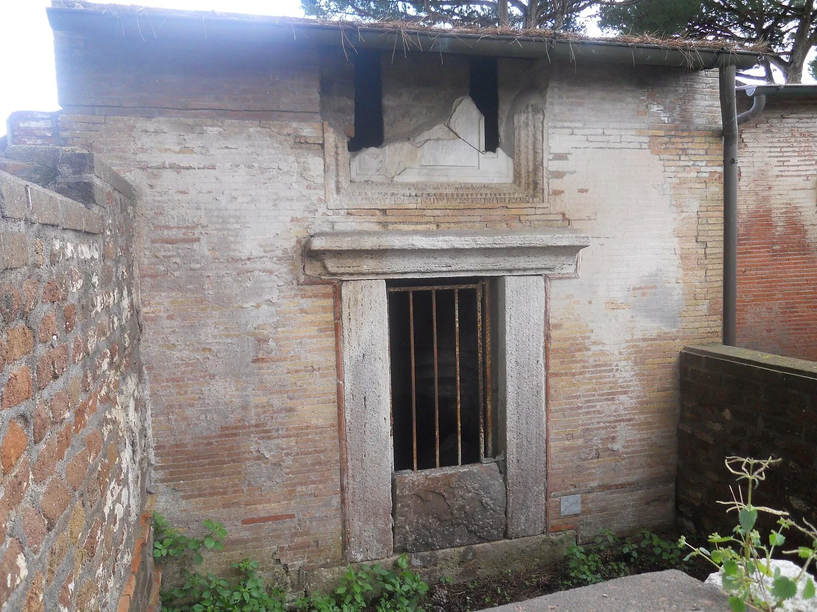 Una tomba della Necropoli di Porto all'Isola Sacra: facciata in mattoni con porta incorniciata da stipiti e architrave in travertino chiusa da un cancello, sopra lo spazio dell'iscrizione in marmo, con un muro in tufo a sinistra