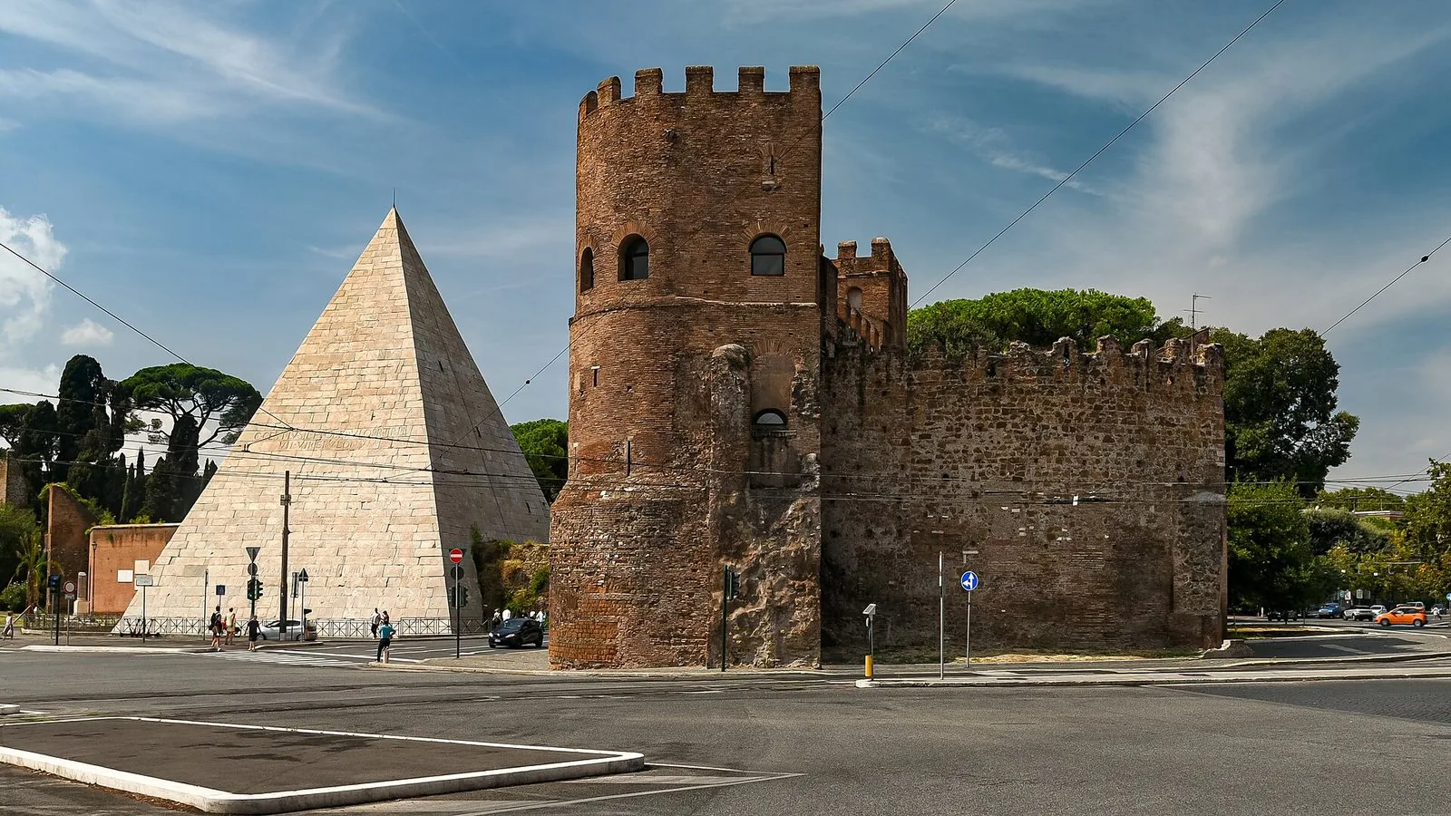 La Piramide Cestia rivestita di marmo bianco accanto a una torre merlata in mattoni delle Mura Aureliane presso Porta San Paolo, con l'incrocio stradale in primo piano e il cielo azzurro