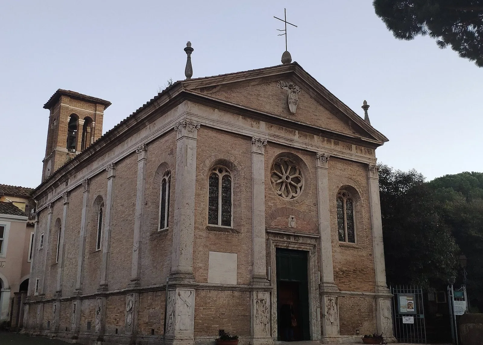 La chiesa di Sant'Aurea nel borgo di Ostia Antica vista di tre quarti: facciata rinascimentale in mattoni e travertino con rosone e timpano, campanile a sinistra e pini sulla destra