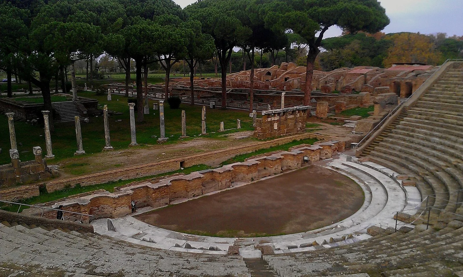 Il teatro romano di Ostia Antica visto dall'alto delle gradinate: la cavea semicircolare, l'orchestra, i resti della scena e, dietro, colonne e rovine in laterizio tra i pini domestici